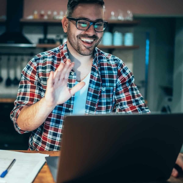 Man at home office waves at the camera on his laptop